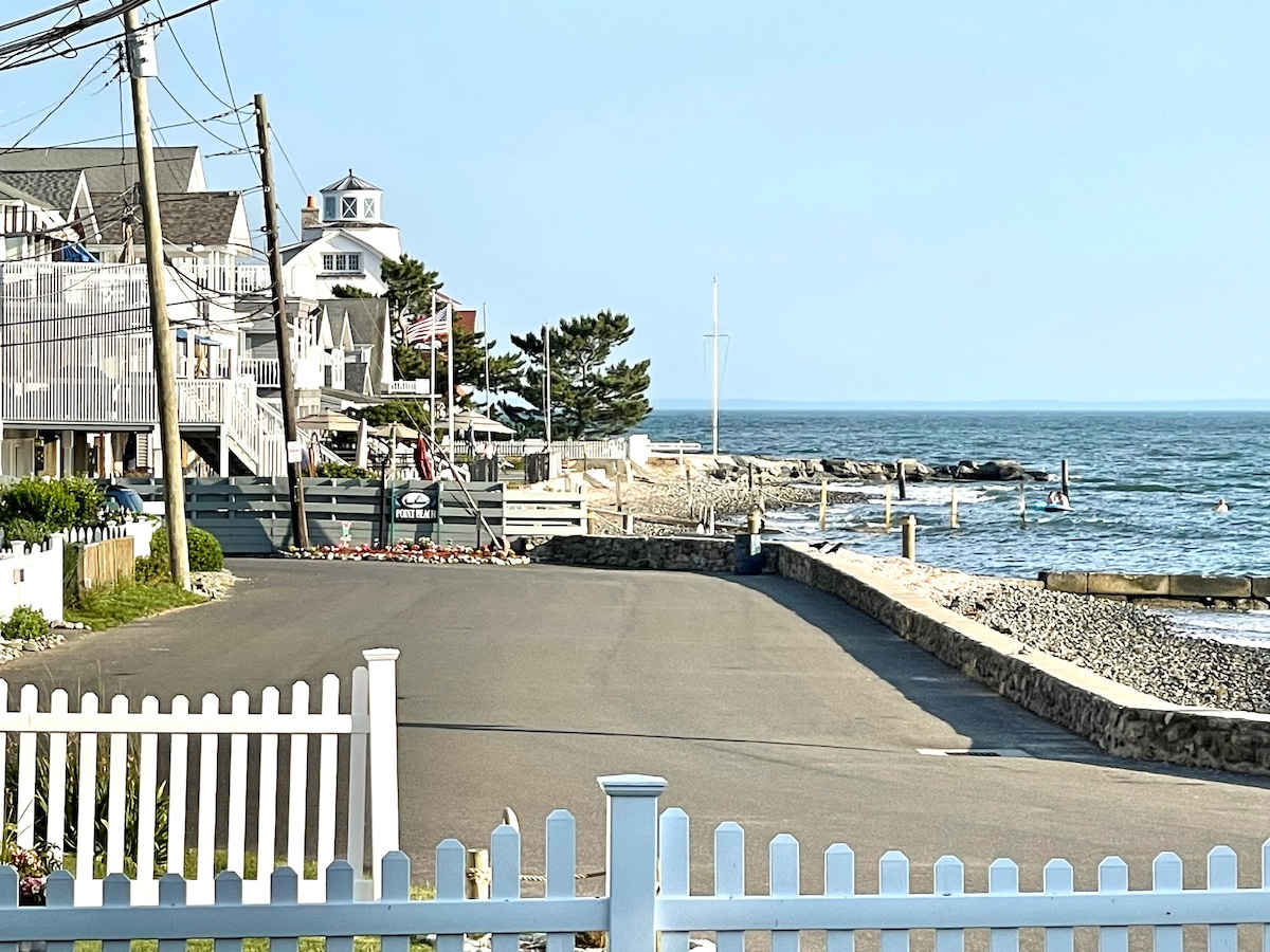 A serene beachfront street is lined with charming coastal homes and cottages. A white picket fence borders the walkway, leading toward the sandy shoreline. The calm ocean waves gently lap at the shore, with people enjoying the water in the distance under a clear blue sky.