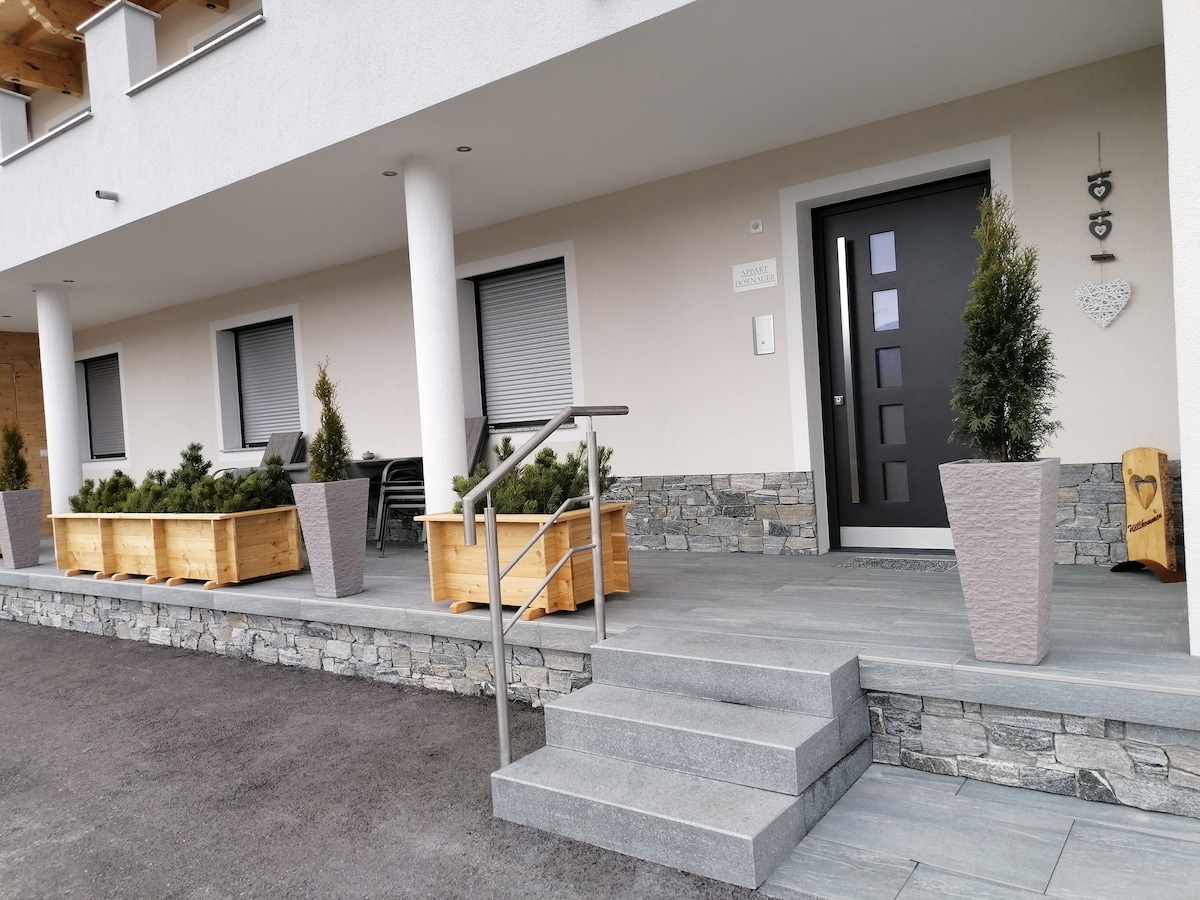 The entrance features a set of concrete steps leading up to a modern front door, framed by decorative planters. Wooden seating is positioned nearby, and stone accents enhance the exterior. The area is well-maintained, with a clean and inviting appearance.
