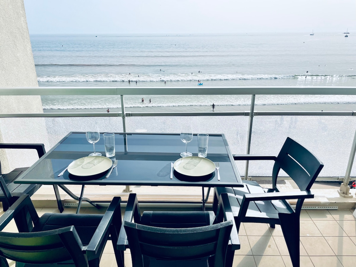 A dining table is set on a private terrace, featuring plates and glasses ready for use. The ocean view is visible through a glass railing, where the shoreline meets the water. Gentle waves can be seen, along with people enjoying the beach.