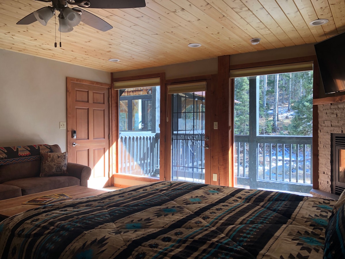 A roomy studio features a queen bed with a patterned blanket, and a sofa positioned nearby. Natural light fills the space through large windows, which offer a view of the outdoor balcony and surrounding forest. A fireplace and wall-mounted television provide additional amenities.