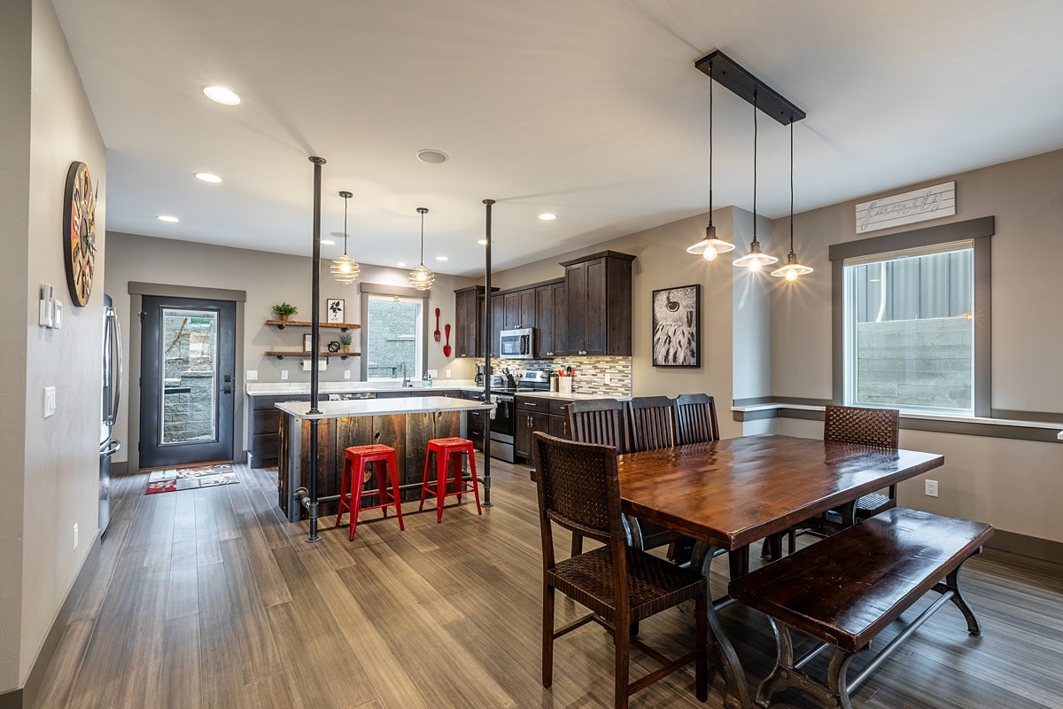 An open kitchen and dining area features modern appliances and dark cabinetry. A large wooden table with seating for six is complemented by red stools at the kitchen island. Large windows provide natural light, enhancing the inviting atmosphere.