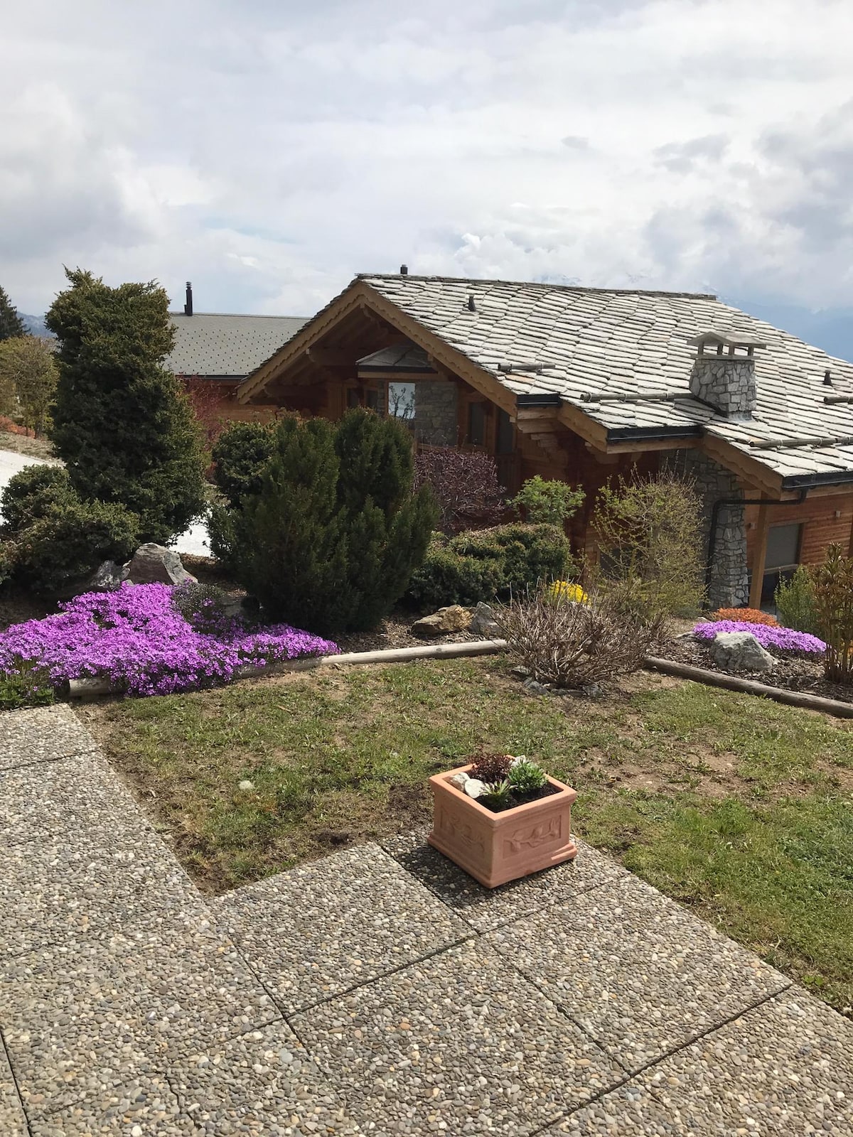 The exterior view of the apartment features a charming cabin-style design with a stone roof. Vibrant purple flowers bloom in the landscaped garden, complemented by lush greenery and a small planter. The pathway is composed of gravel and stone, enhancing accessibility.