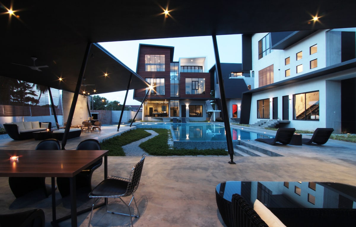 An outdoor area is shown, featuring a modern swimming pool surrounded by well-manicured grass. Stylish black lounge chairs and a wooden dining table are situated under a sleek roof structure, creating a cozy space for relaxation and dining. The contemporary building is visible in the background.