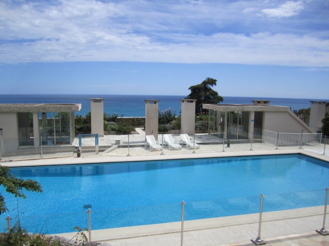 A clear blue swimming pool is surrounded by a spacious terrace, featuring several lounge chairs facing the ocean. The backdrop includes landscaped greenery and a view of the sea under a partly cloudy sky, creating a serene coastal environment.