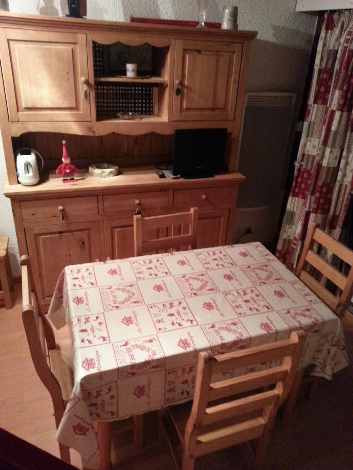 A wooden dining table is set with a patterned tablecloth, surrounded by four wooden chairs. A hutch displays decorative items and contains a kettle and a small television. Soft lighting enhances the warm ambiance of the room.