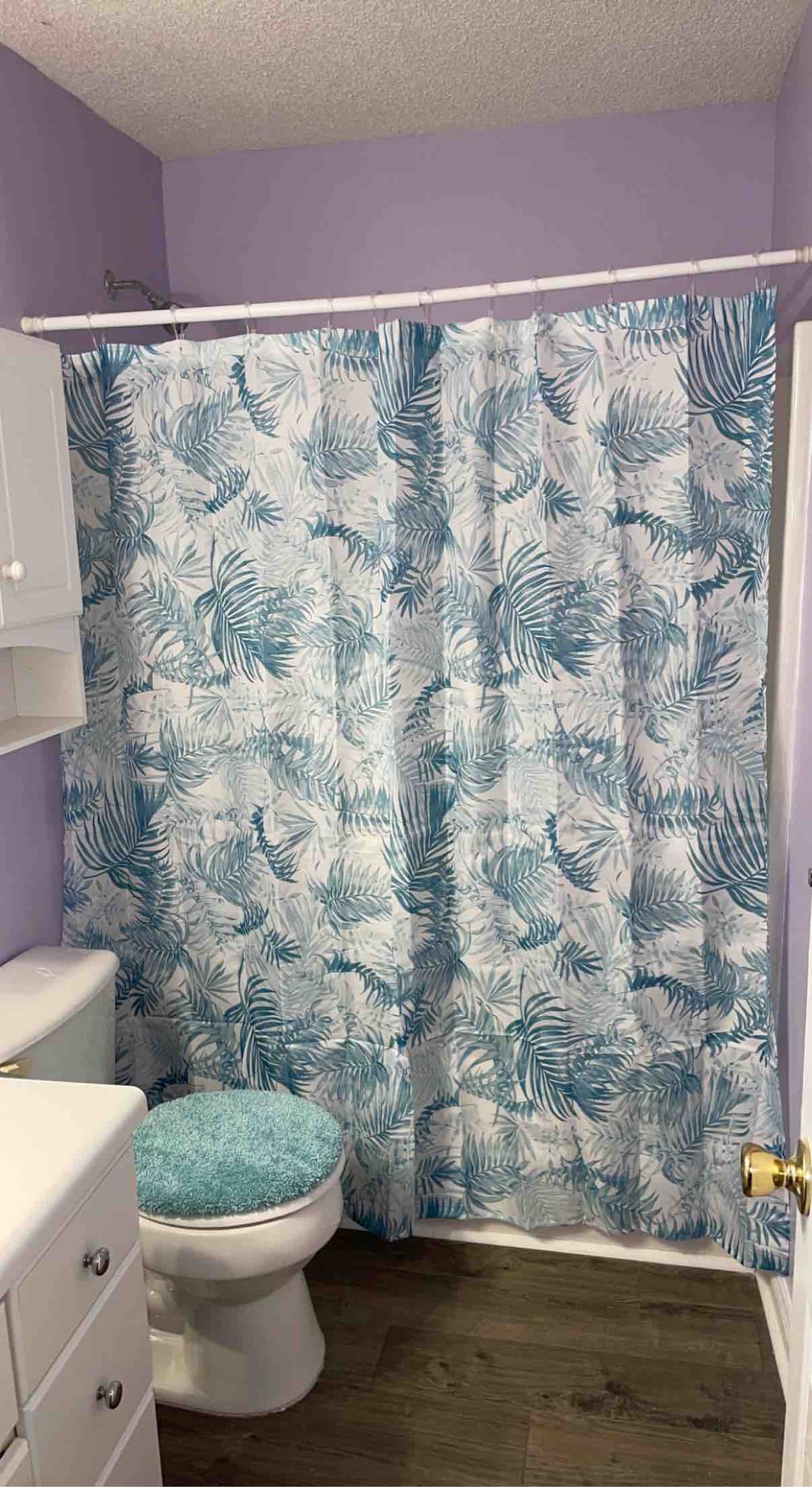 A bathroom features a soft blue and white tropical-themed shower curtain, creating a calming visual. A matching bath mat sits in front of the commode. Light purple walls complement the white cabinetry and sink, while a neutral floor adds to the serene ambiance.