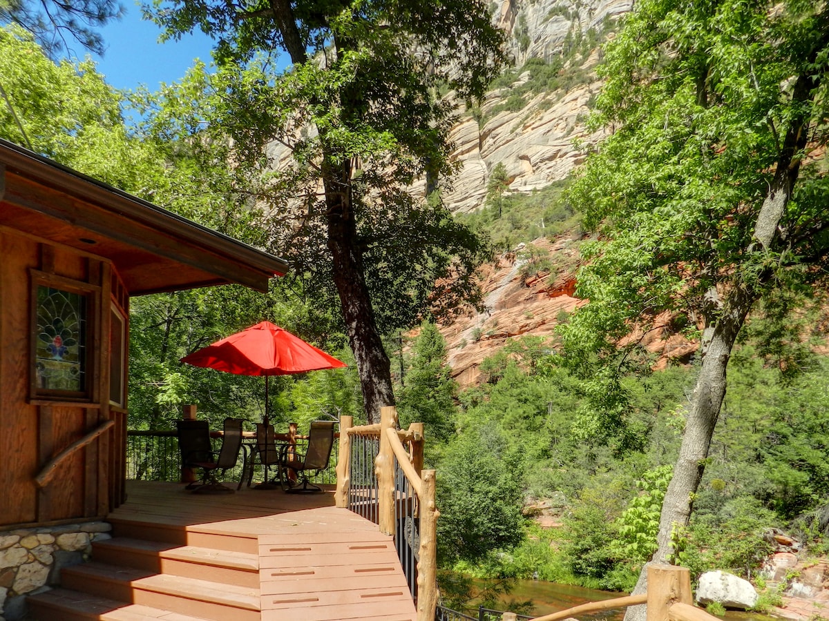 A covered deck area features a red umbrella over a dining table, surrounded by chairs. The backdrop showcases towering red rock cliffs and lush greenery, enhancing the natural setting. Steps lead down to the creek, providing easy access to the serene outdoor environment.