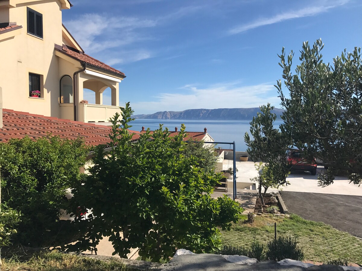 The image showcases a serene view of a coastal area, featuring a well-maintained building with terracotta roofing. A green garden with shrubs and trees is present in the foreground, while the tranquil blue sea and distant cliffs are visible beyond.