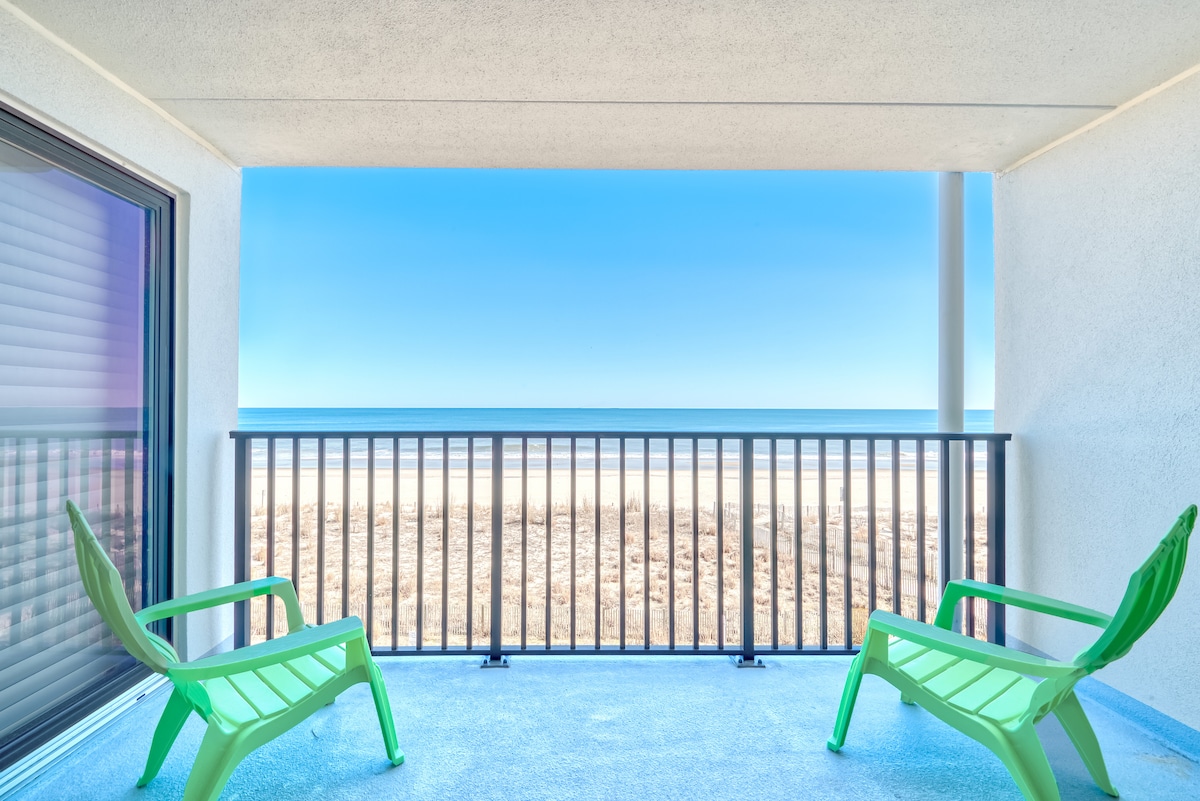 A private balcony showcases two green lounge chairs facing the ocean, with a clear blue sky and sandy beach visible in the background. The railing adds a sense of safety while maintaining an unobstructed view of the serene seaside landscape.