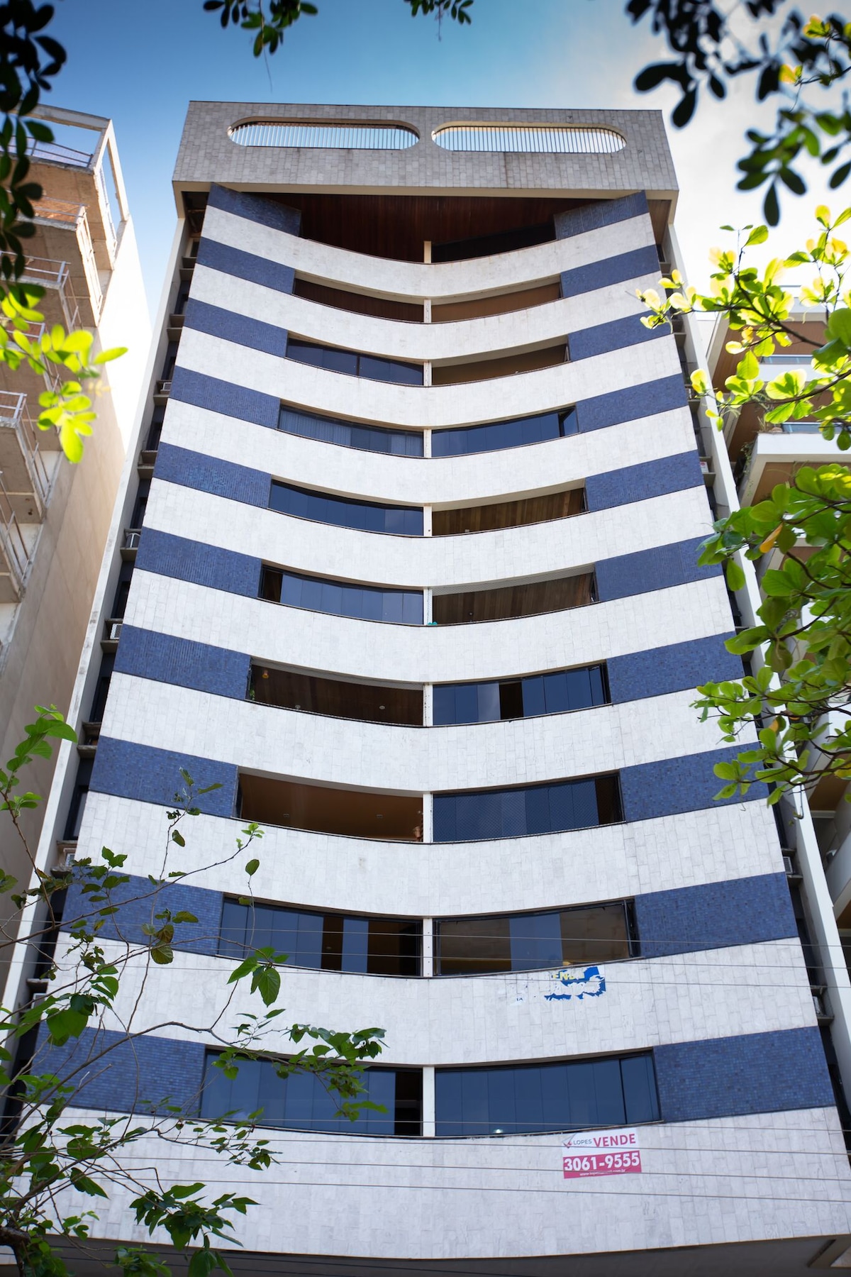 The exterior of a modern multi-story building features alternating blue and white striped facades. Large windows span each floor, providing a bright appearance. Green foliage surrounds the base, subtly framing the structure while a sign indicates that the property is for sale.
