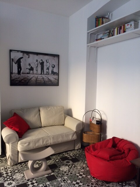 A cozy seating area is presented with a light-colored sofa adorned with a red cushion. Next to it, a round side table stands on patterned tiled flooring. A bright red bean bag adds a pop of color, while a wall-mounted shelf displays a few books above.