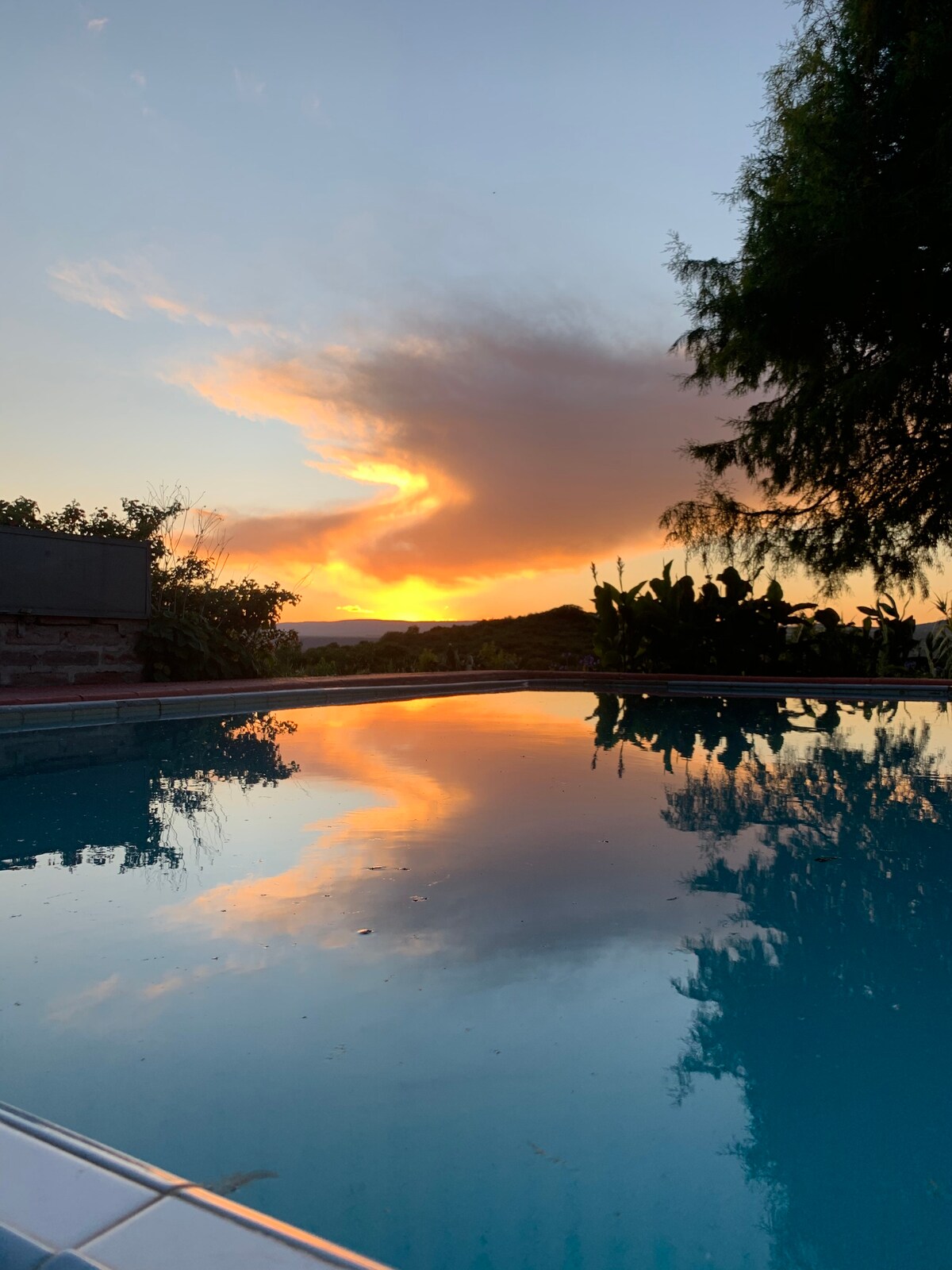 A serene swimming pool reflects vibrant hues of orange and pink from a sunset sky, surrounded by lush greenery. The tranquil water creates a peaceful ambiance, enhancing the natural beauty of the landscape beyond.