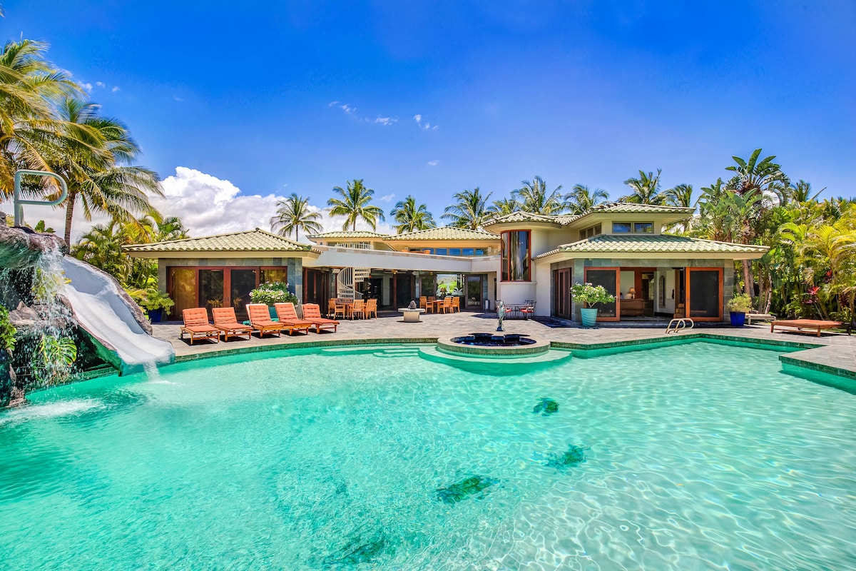 The spacious pool area is highlighted by a saltwater pool with a waterfall and a water slide. Sun loungers are arranged around the pool, surrounded by lush landscaping and palm trees. A clear blue sky complements the tropical setting of the property.