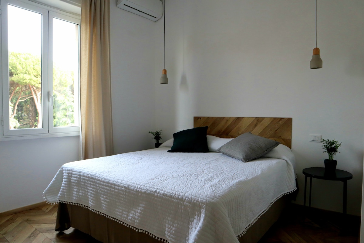 A well-lit bedroom features a spacious bed with a textured headboard. Two hanging light fixtures provide soft illumination. Green plants are placed on a small bedside table, next to the bed, while natural light enters through a large window, framed by light curtains.