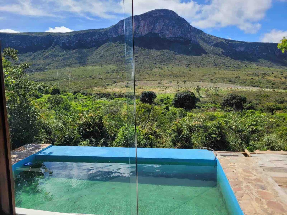 A refreshing pool is depicted, surrounded by natural greenery, with a panoramic view of a large mountain in the distance. The crystal-clear water reflects the sky, enhancing the serene atmosphere of the rural setting.