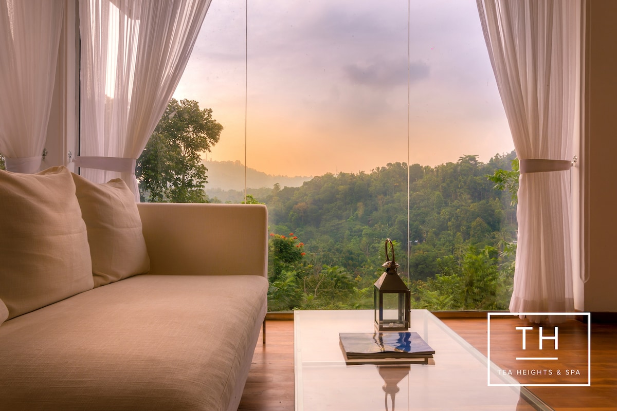 A spacious seating area is highlighted by a comfortable sofa positioned next to a clear glass window, providing an expansive view of the lush green landscape and hills. A lantern rests on a sleek coffee table, gently illuminating the space in the soft light of dawn.