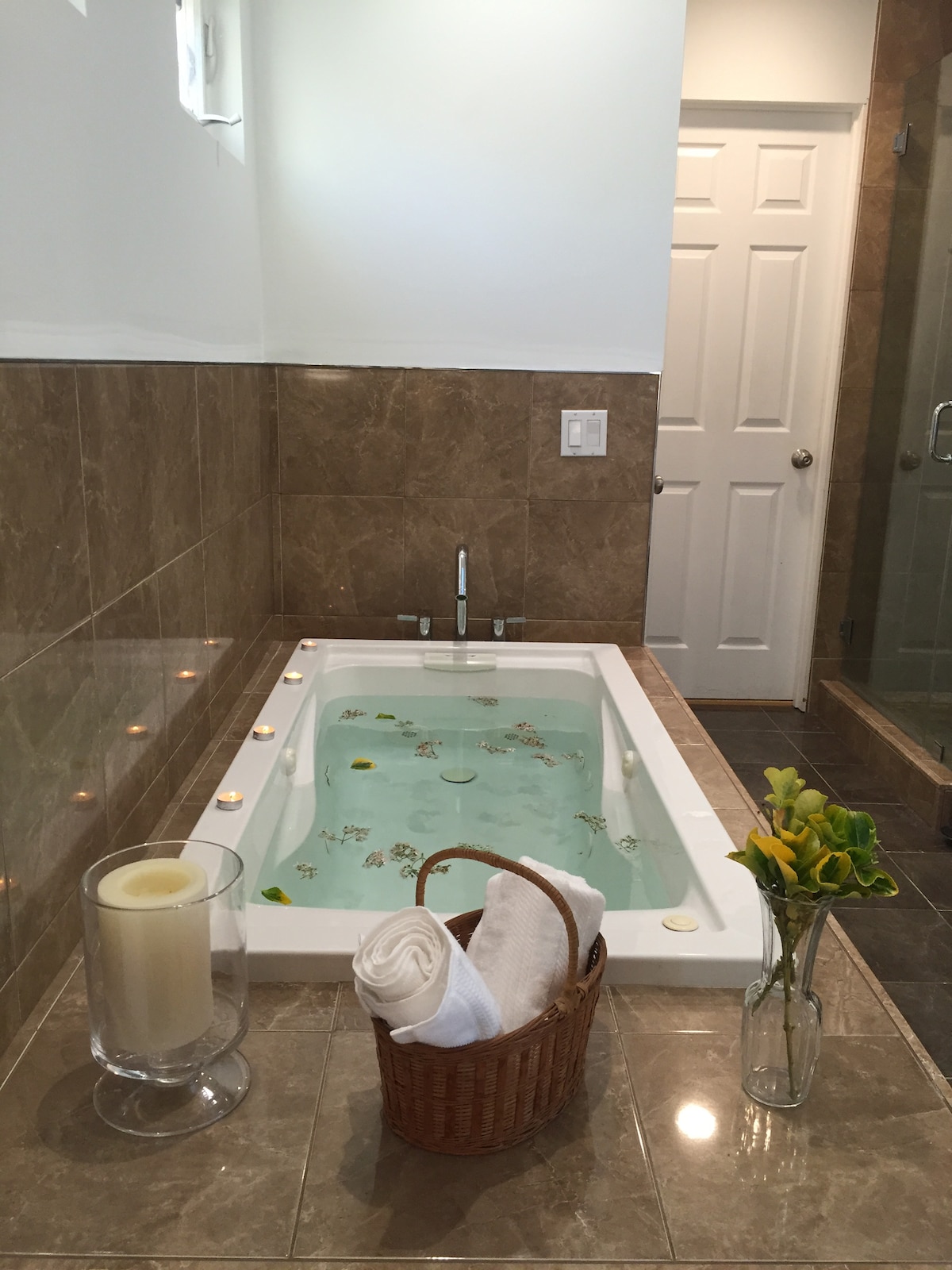 A relaxing bathroom features a jetted tub filled with water and adorned with floating petals. Soft lighting surrounds the tub, emphasizing the elegant tiled walls. A wicker basket holds rolled towels, while a simple floral arrangement adds a natural touch on the countertop.