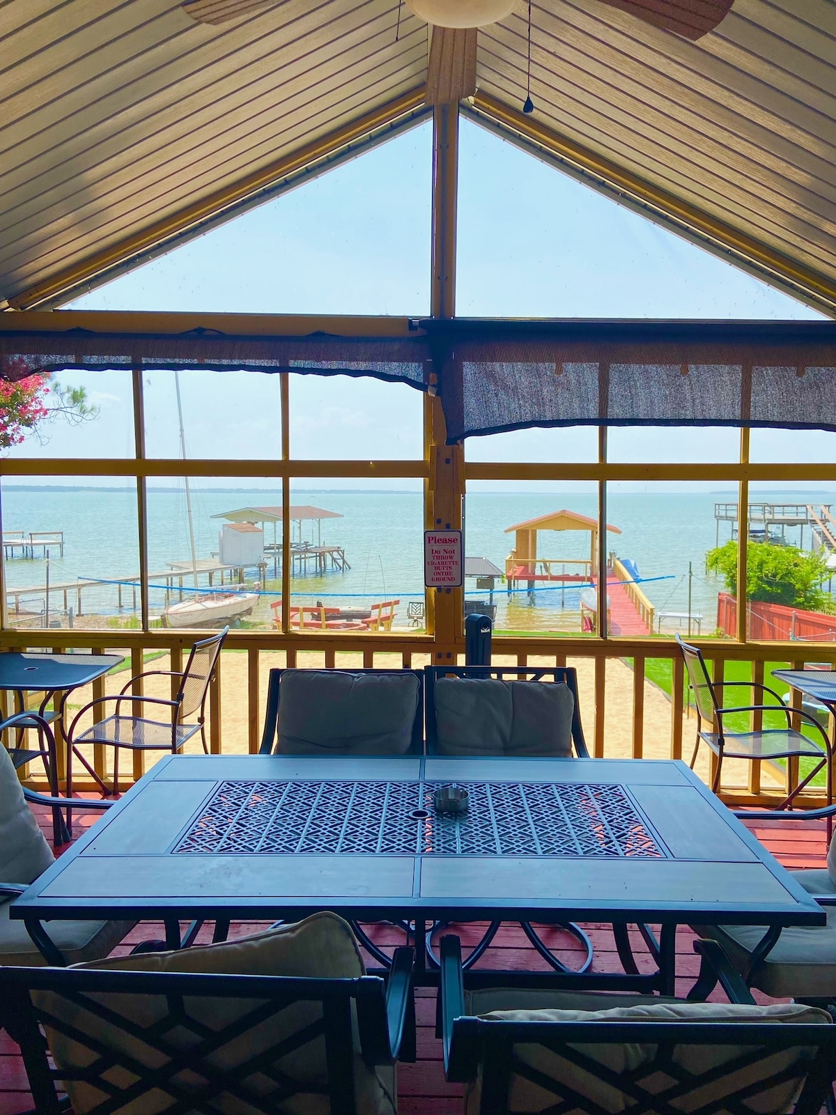 A spacious seating area is framed by large windows, offering a panoramic view of the water. Comfortable chairs surround a table, creating an inviting space for relaxation and socializing. The bright blue lake and outdoor area are visible through the glass, enhancing the serene atmosphere.