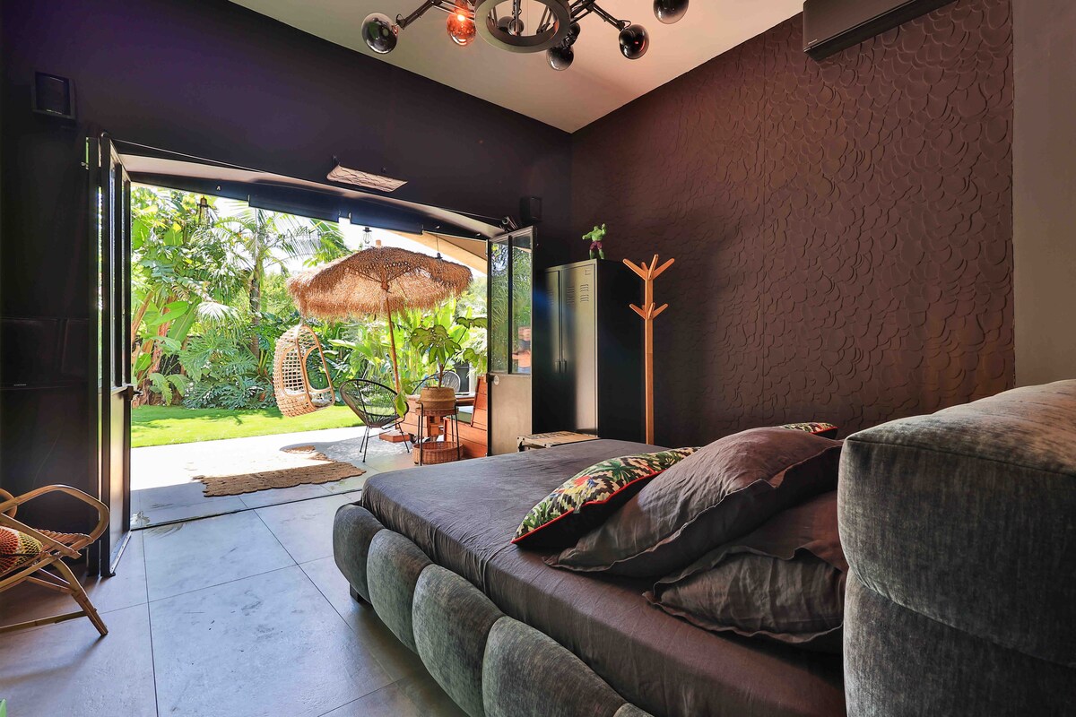 A spacious bedroom features a comfortable bedding arrangement with decorative pillows. Large glass doors open to a lush garden area, showcasing a seating arrangement and a hanging chair under a shaded umbrella. Natural light enhances the inviting ambiance.