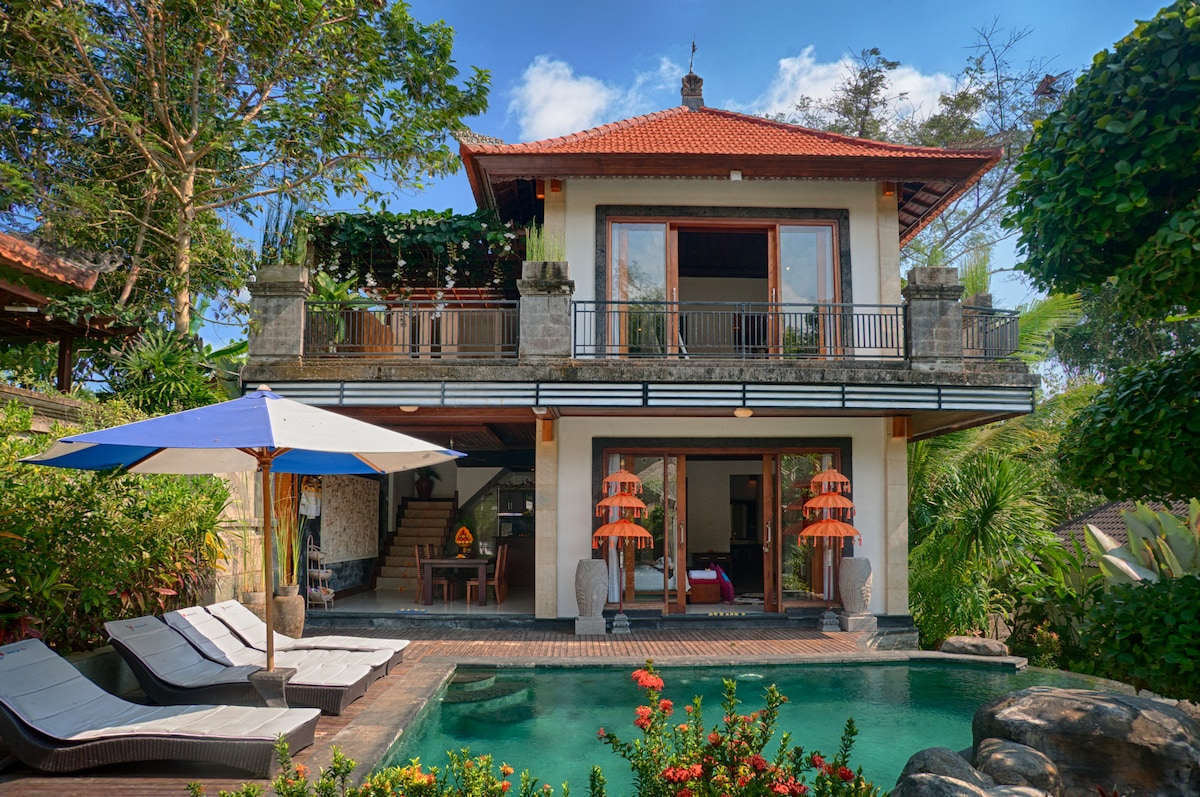 The villa showcases a two-story structure with a traditional Balinese roof, surrounded by lush greenery. A private pool is visible in the foreground, framed by lounge chairs and colorful flowers. Large glass doors provide access to a spacious balcony on the upper floor.