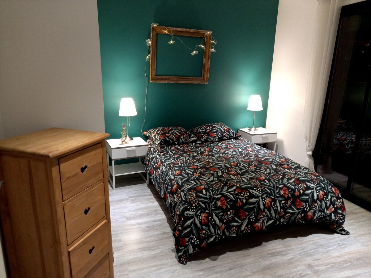 A cozy bedroom is presented with a dark floral-patterned bedspread covering a spacious bed. Two bedside tables with lamps are positioned on either side. A wooden chest of drawers stands against one wall, while a large mirror adorns the green feature wall, complemented by subtle string lights.