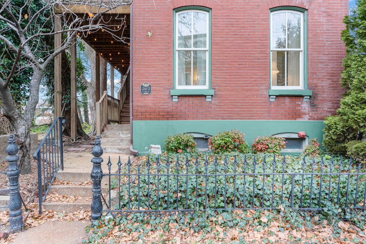 A charming Victorian duplex is featured, showcasing a red brick exterior with contrasting green accents. A wrought iron fence lines the front, enclosing a small garden filled with ivy and fallen leaves. Decorative string lights can be seen along the entrance leading to the front door.