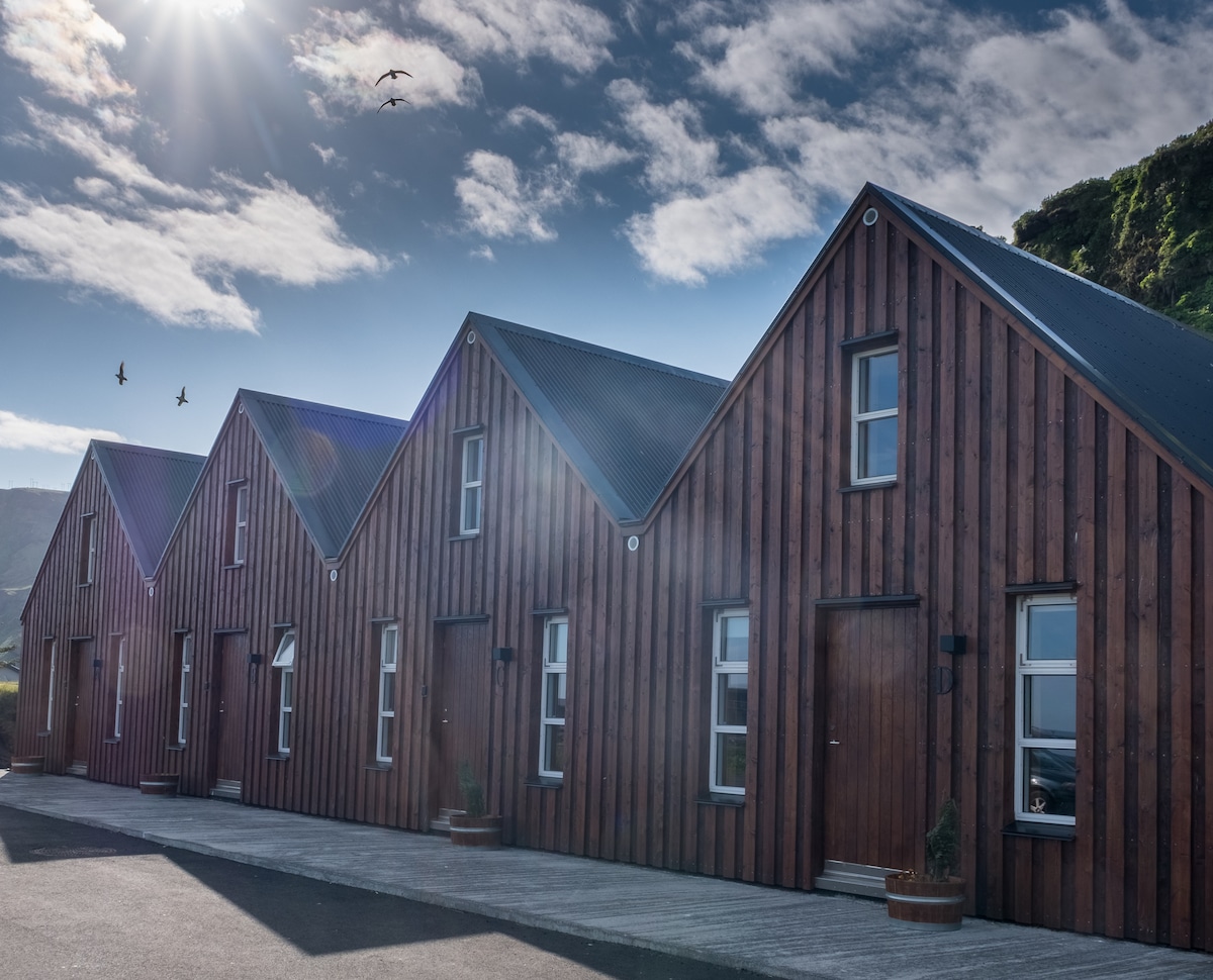 A row of newly constructed cabins features a modern wooden exterior with a sloped roof. Sunlight highlights the warm tones of the wood, while clear skies and distant birds create a serene backdrop. Each cabin is equipped with a private entrance.