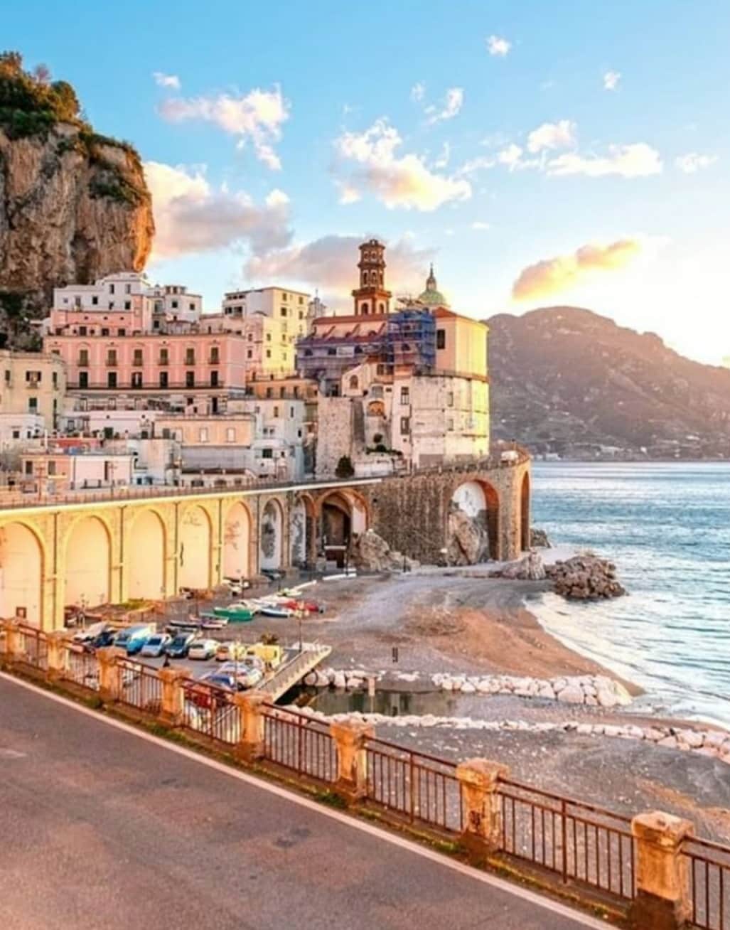 A scenic view captures colorful buildings on the cliffside, nestled by the tranquil waters. The coastline is lined with boats and a sandy shore, framed by majestic mountains under a warm sunset sky. This serene setting exemplifies the charm of Atrani, along the Amalfi Coast.