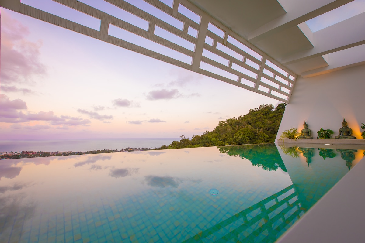 An infinity pool stretches out seamlessly with reflections of the sky and surrounding greenery. The pool area is framed by unique architectural elements above, allowing natural light to filter through. The tranquil atmosphere is enhanced by the soft colors of sunset as they blend into the horizon.