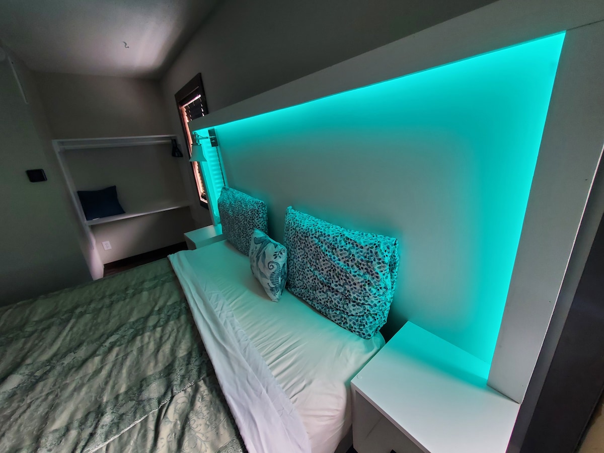 The studio features a modern sleeping area with two pillows resting against a backlit headboard, emitting a soft turquoise glow. A spacious bed is positioned in front, flanked by minimalistic nightstands, while a shelving unit is placed to one side.