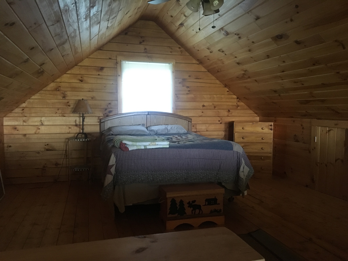 The loft sleeping area features wooden walls and flooring, with a spacious bed dressed in assorted linens. A small bedside lamp is positioned next to the bed, and a decorative chest sits at the foot, providing both function and warmth to the rustic setting.