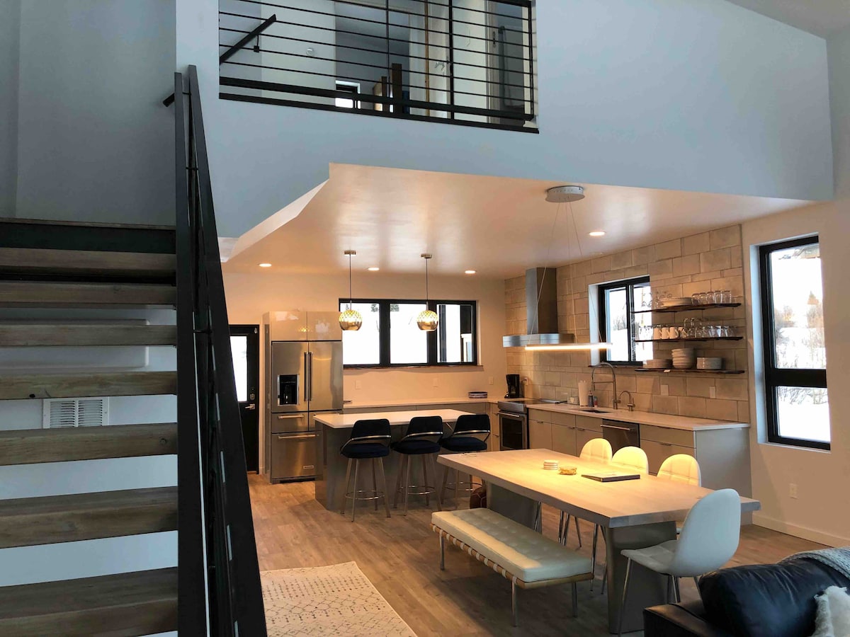 An open-concept living space is showcased, featuring a modern kitchen with stainless steel appliances and ample counter space. A dining table with seating for eight sits beside a light-colored bench. A staircase leads to an upper level, framed by sleek railings.