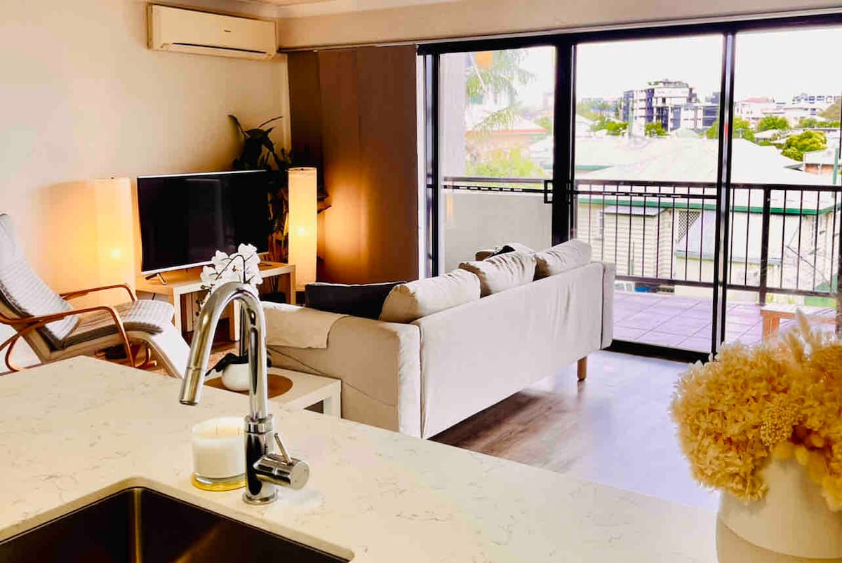 The airy living area is furnished with a light-colored sofa and rattan chair, complemented by a mounted television. A view of the balcony is visible through sliding glass doors, which allow natural light to fill the space. A modern kitchen island with a sink is positioned in the foreground.