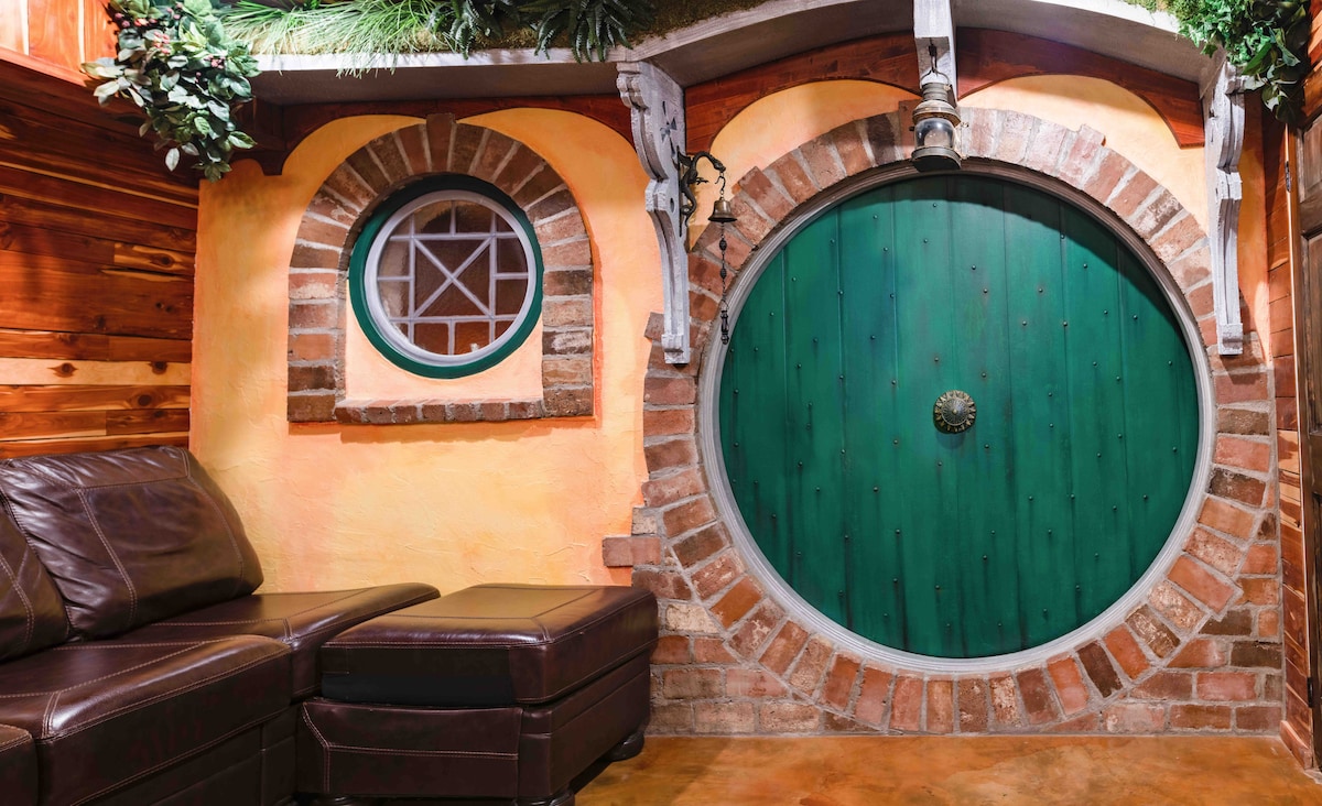 A charming brick Hobbit door with a circular design is framed by rustic stonework. An adjacent round window features a grid pattern, complementing the door's design. A leather couch sits nearby, inviting relaxation in a cozy, woodland-themed space.