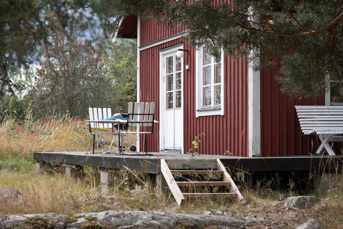 The exterior of the cottage features a red wood façade with white trim. A small staircase leads to a deck, where outdoor furniture, including tables and chairs, is arranged. Surrounding grass and natural vegetation provide a relaxed setting near the entrance.