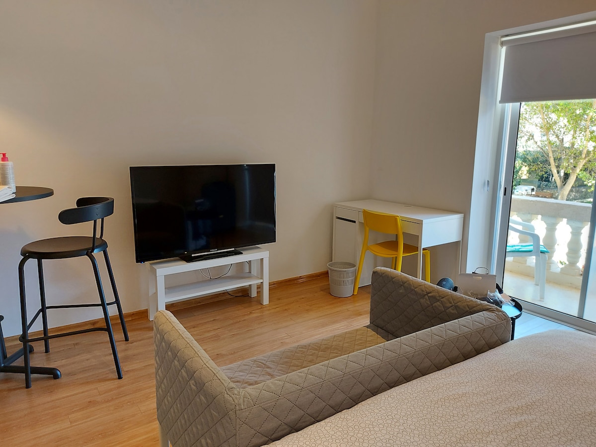 A modern studio is depicted, featuring a spacious bed with a textured cover. A television is positioned on a white console, while a small dining table with high stools is seen nearby. A desk with a yellow chair is also present, alongside a sliding door leading to a balcony.