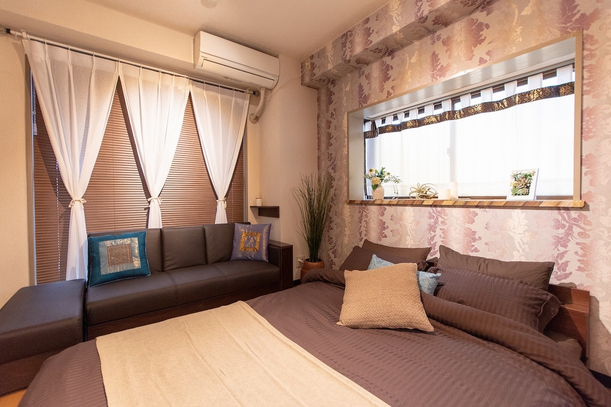 A cozy sleeping area features a spacious bed dressed in dark linens, complemented by decorative cushions. A sofa with colorful pillows is positioned nearby, while light filters in through sheer curtains, illuminating a small window with potted plants.