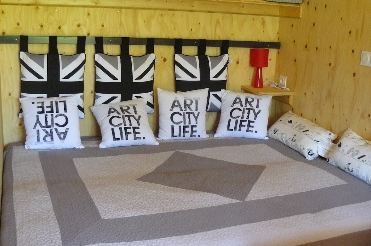 A comfortable sleeping area is presented, featuring a spacious bed adorned with a neutral-toned quilt. The headboard is lined with black cushions displaying a graphic design, complemented by decorative pillows with text. A small bedside table with a lamp is positioned nearby.
