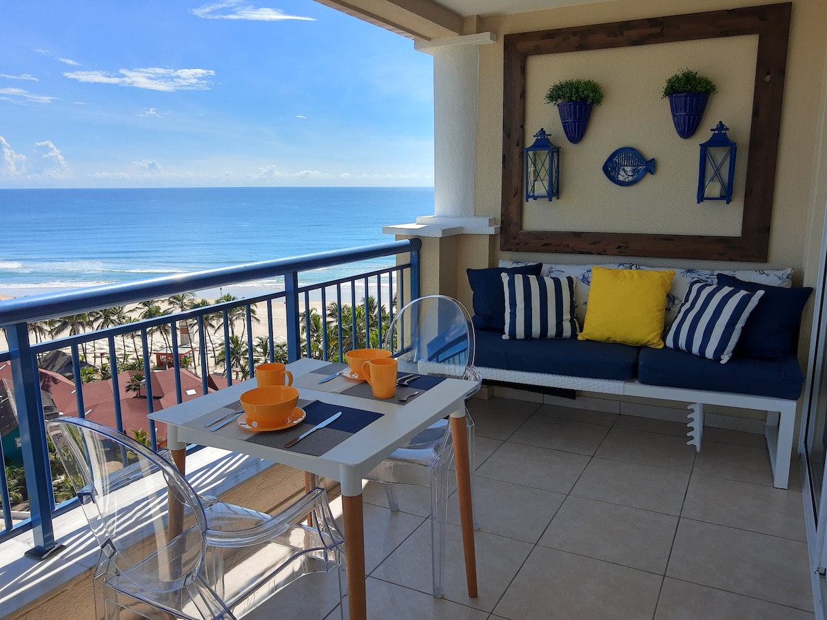 A cozy balcony area is featured, offering ocean views. A small table is set with two orange cups and a yellow accent pillow complements the cushioned seating. Blue and white decor elements create a calming environment, with the beach and palm trees visible in the background.