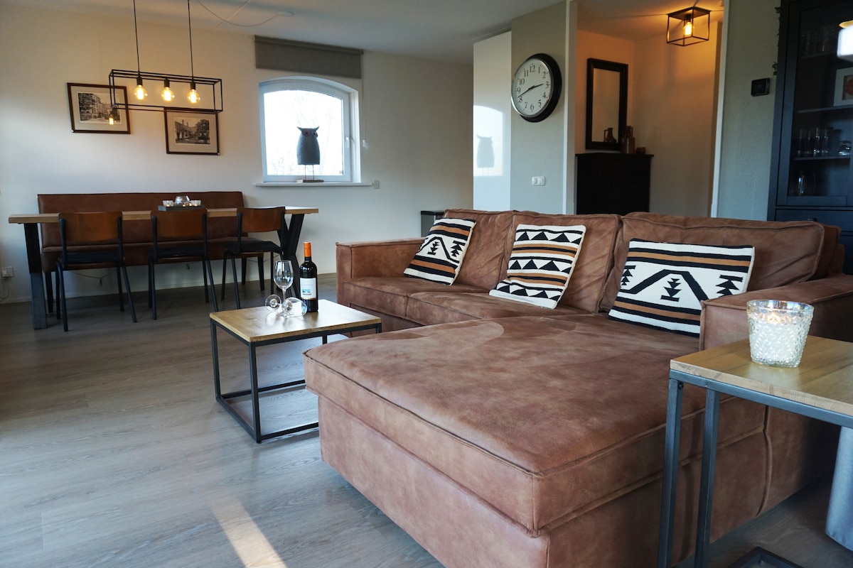 A modern living area features a spacious brown sofa with patterned cushions, accompanied by a coffee table. Dining seating for four is visible in the background, with pendant lighting above. A wall clock and decorative items add subtle detail to the light-filled space.