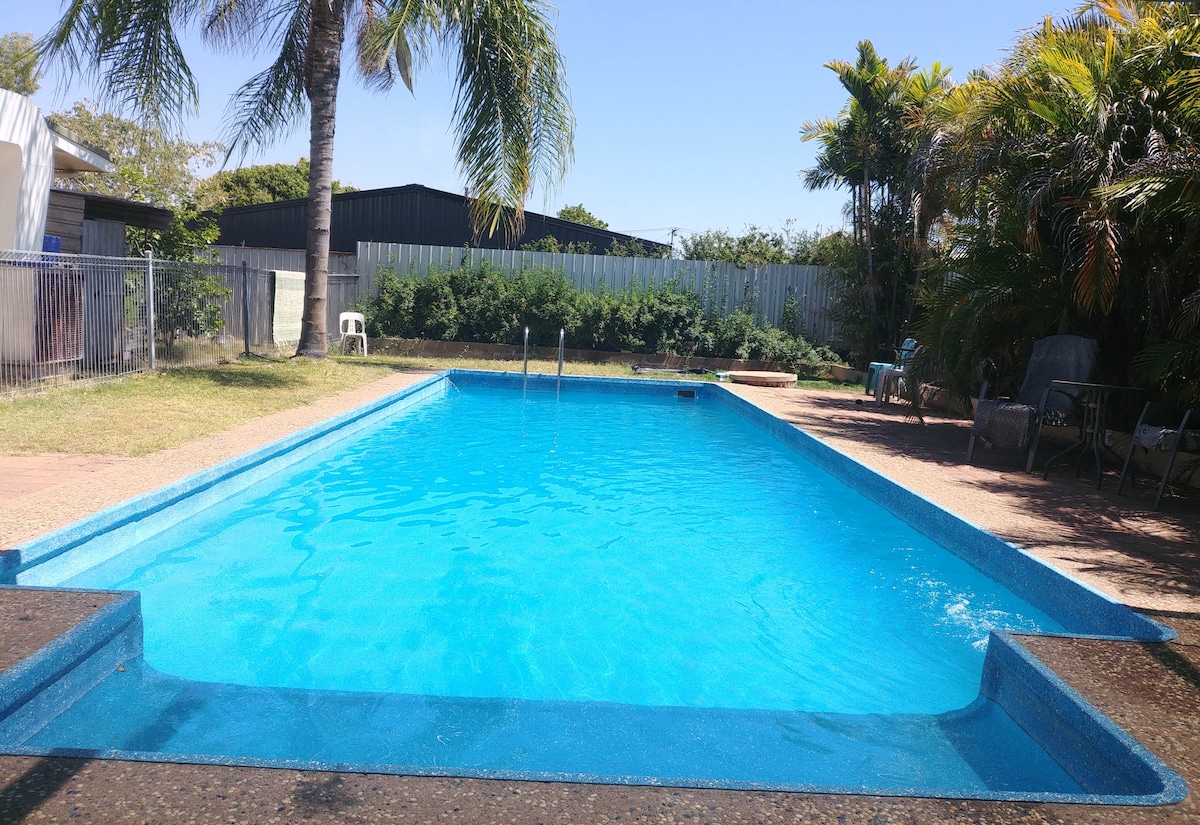 A rectangular pool with clear blue water is framed by a stone deck and surrounded by greenery and palm trees. A few lounge chairs are positioned nearby, providing a pleasant space for relaxation. The open sky and sunlight contribute to a welcoming outdoor area.