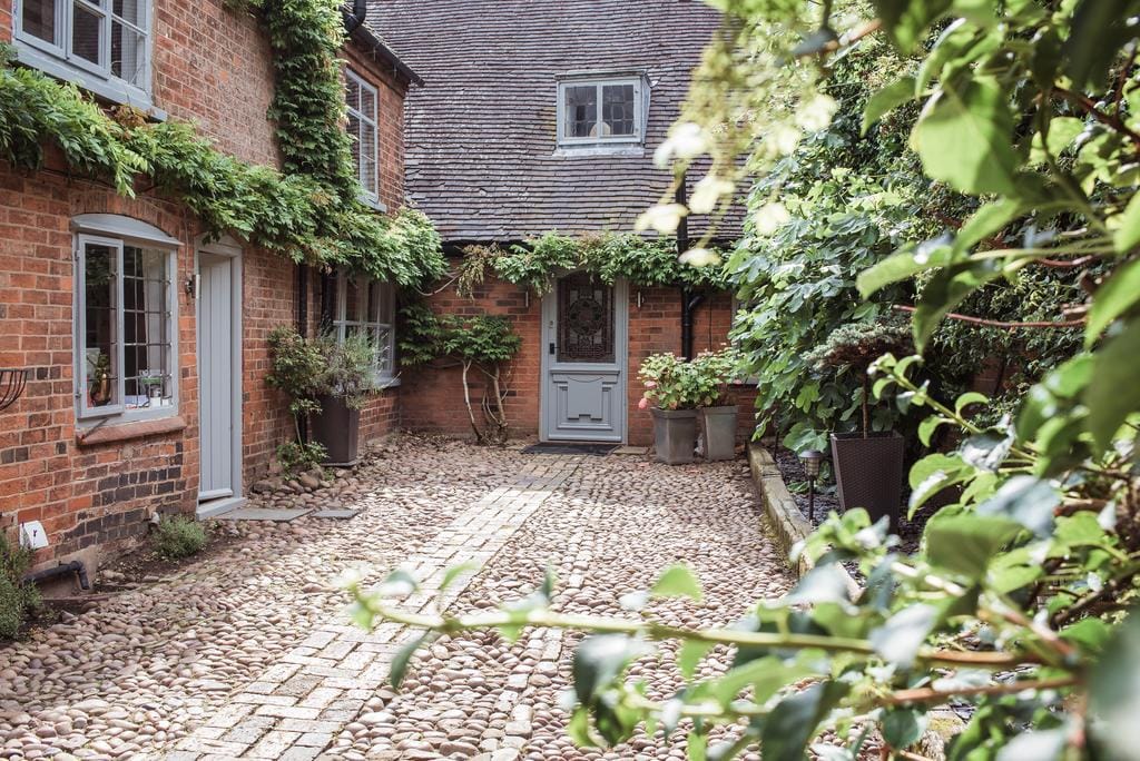 A cobbled courtyard is framed by greenery, showcasing the natural stone path leading to a grey door. The warm brick walls of the surrounding buildings are softened by climbing plants, creating a serene entrance to the property.