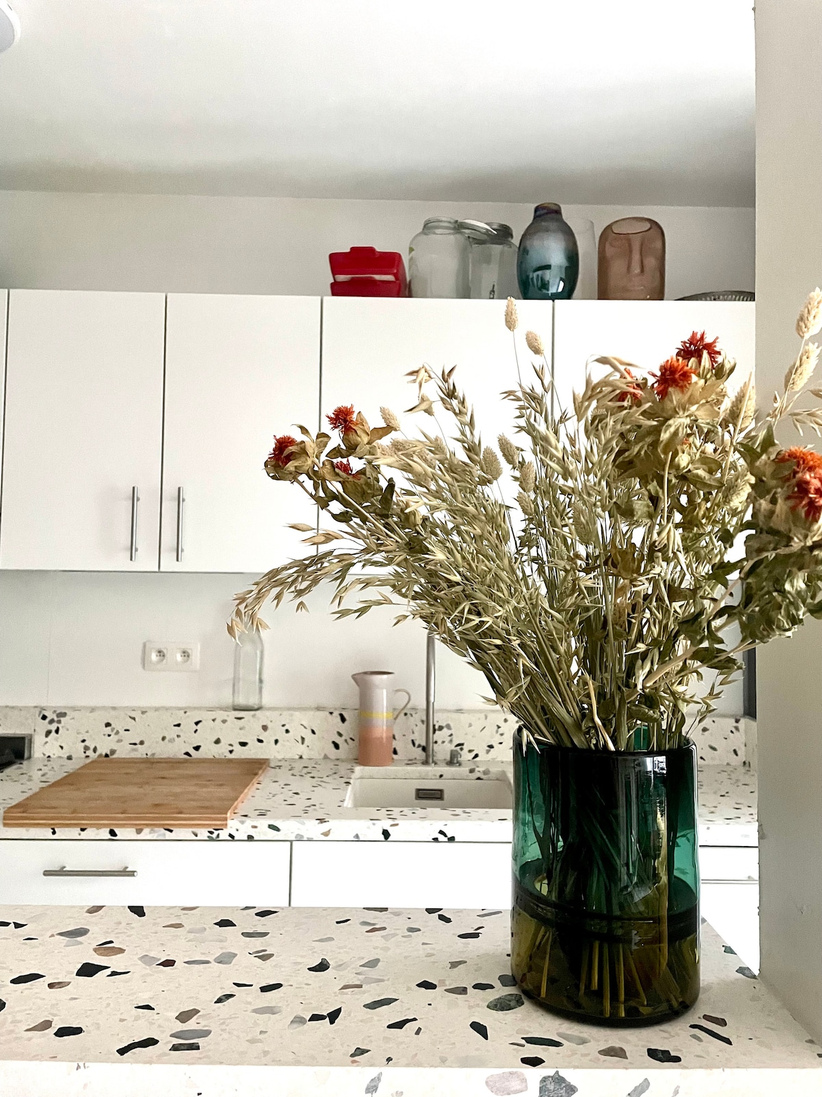 A kitchen is highlighted by a textured countertop featuring a mix of light and dark hues. A glass vase filled with dried flowers stands prominently, adding a natural element to the space. Minimalistic white cabinets complement the contemporary design.