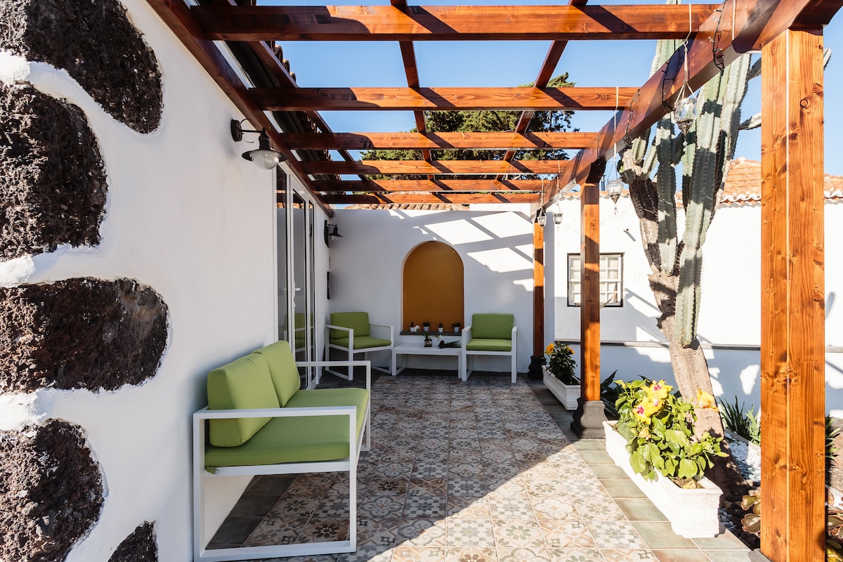 An outdoor seating area is presented under a wooden pergola, featuring white modern furniture with green cushions. Framed by stone walls and potted plants, a textured tiled floor adds character to the space while natural light enhances the inviting atmosphere.