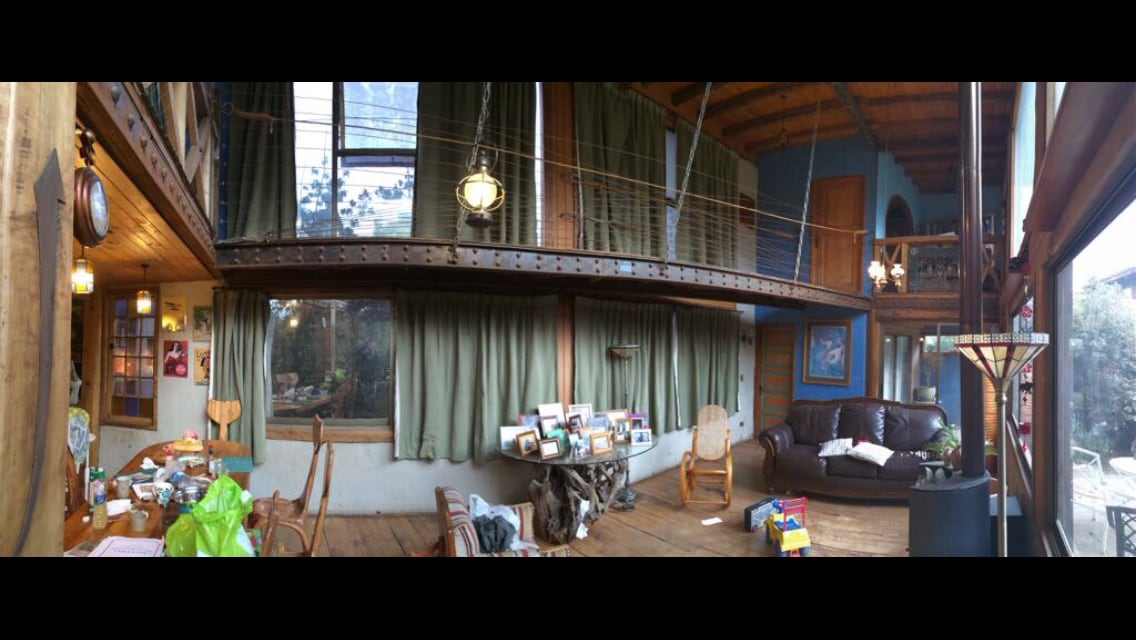 An open living space showcases a blend of rustic charm and modern comfort. Wooden beams and curtains frame large windows, while a comfortable sofa and a wooden table with framed photographs create an inviting gathering area. Various decorative items add personal touches throughout the room.