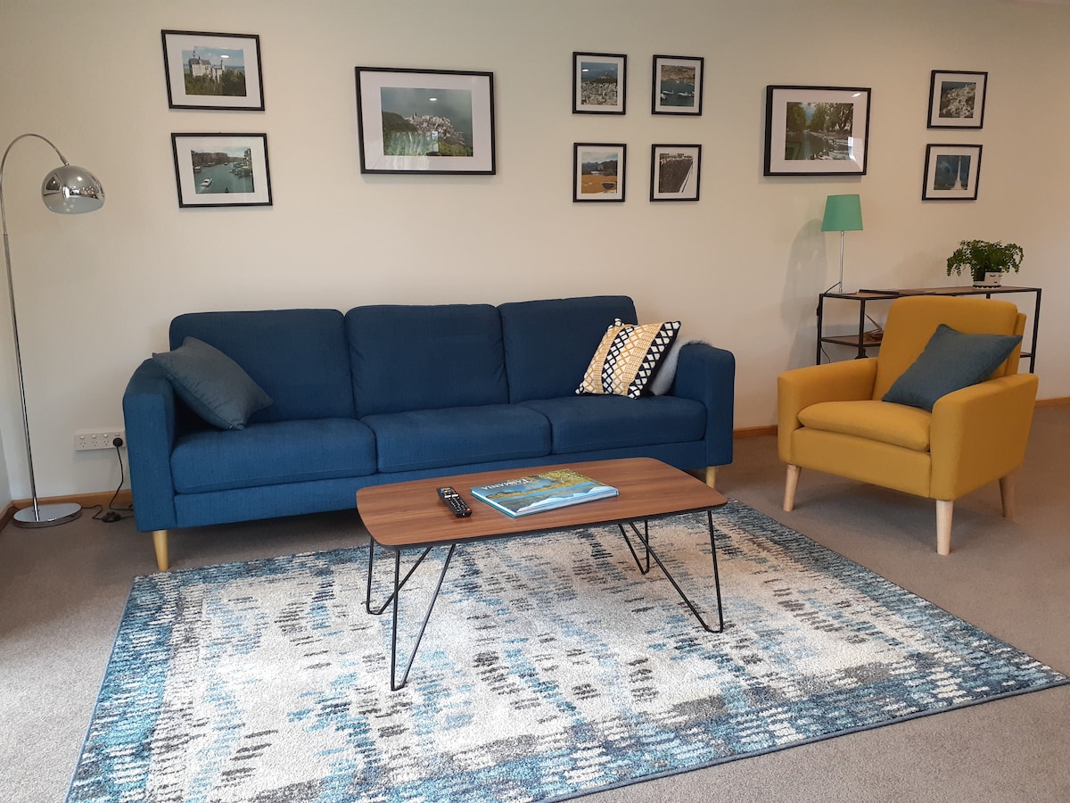 A comfortable lounge area is presented, featuring a blue sofa and a mustard yellow armchair. A coffee table is centered on a patterned rug. Framed photographs adorn the walls, providing a personal touch to the space. Soft lighting is provided by a floor lamp.