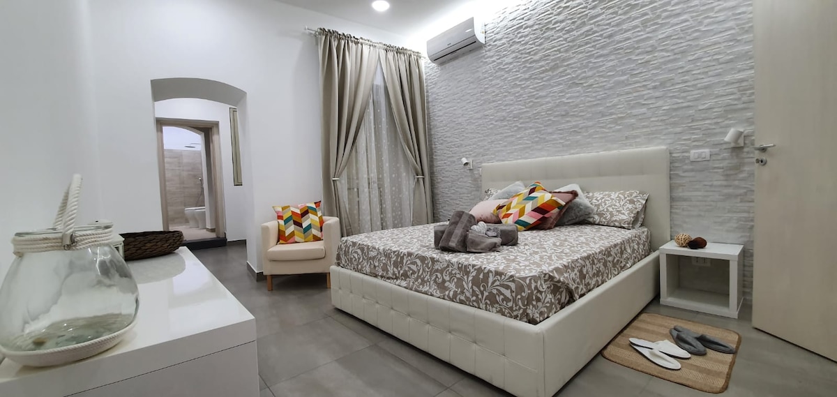A spacious bedroom features a large bed adorned with a patterned coverlet and decorative pillows. A comfortable chair sits nearby, and a light rug lies on the floor. Soft natural light streams through the window, which is dressed with sheer curtains.