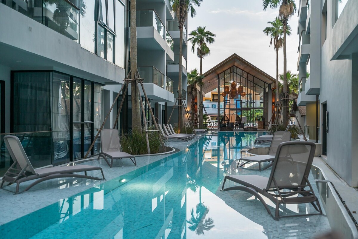 A tranquil swimming pool is framed by palm trees, with loungers positioned alongside the water. The pool area reflects a modern design, featuring clear blue water that invites relaxation. A clubhouse with large windows is visible in the background, providing a welcoming communal space.