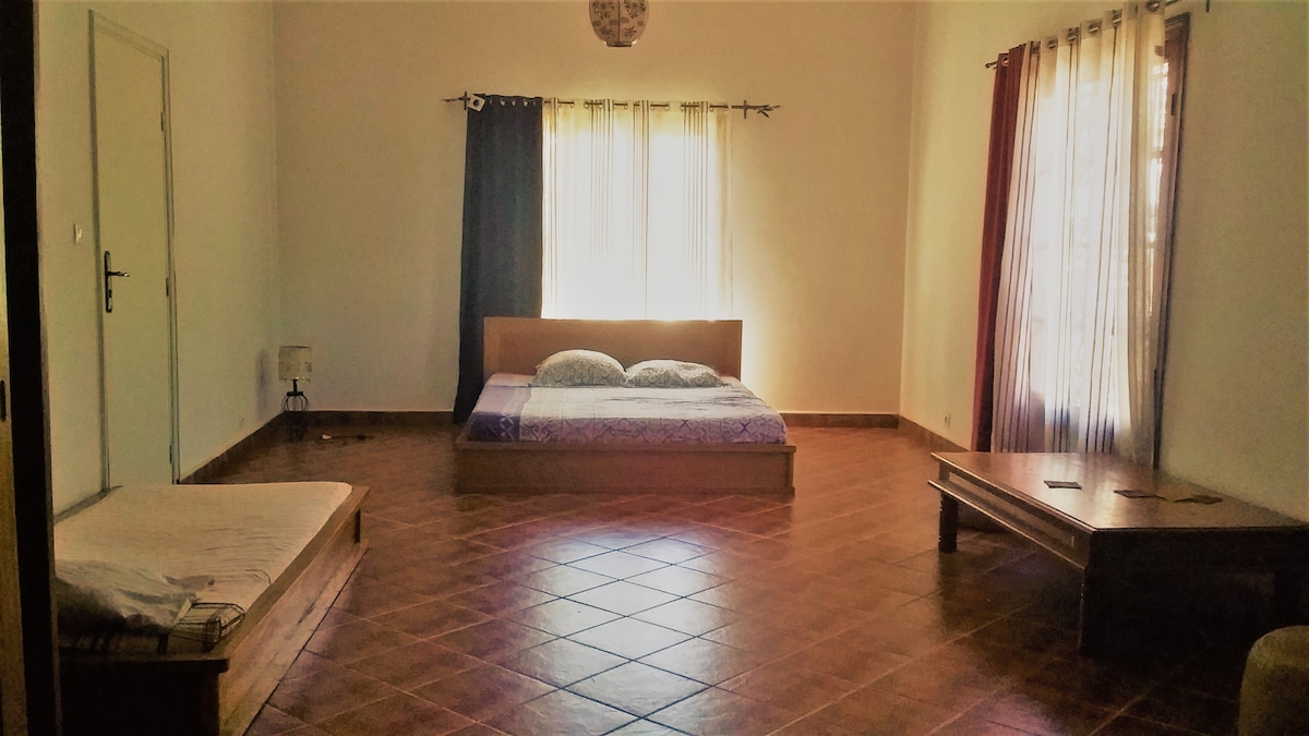 A spacious room is highlighted by a large bed with a patterned blanket. Natural light filters through large windows, adorned with light-colored curtains. A wooden coffee table and a separate mattress are positioned on the tiled floor, contributing to the room's open design.