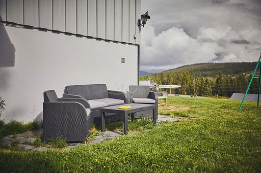 An outdoor seating area is arranged on a paved stone surface, featuring a dark wicker sofa and coffee table. In the background, lush green grass extends towards a wooden table, with a swing set and distant forested hills under a partly cloudy sky.